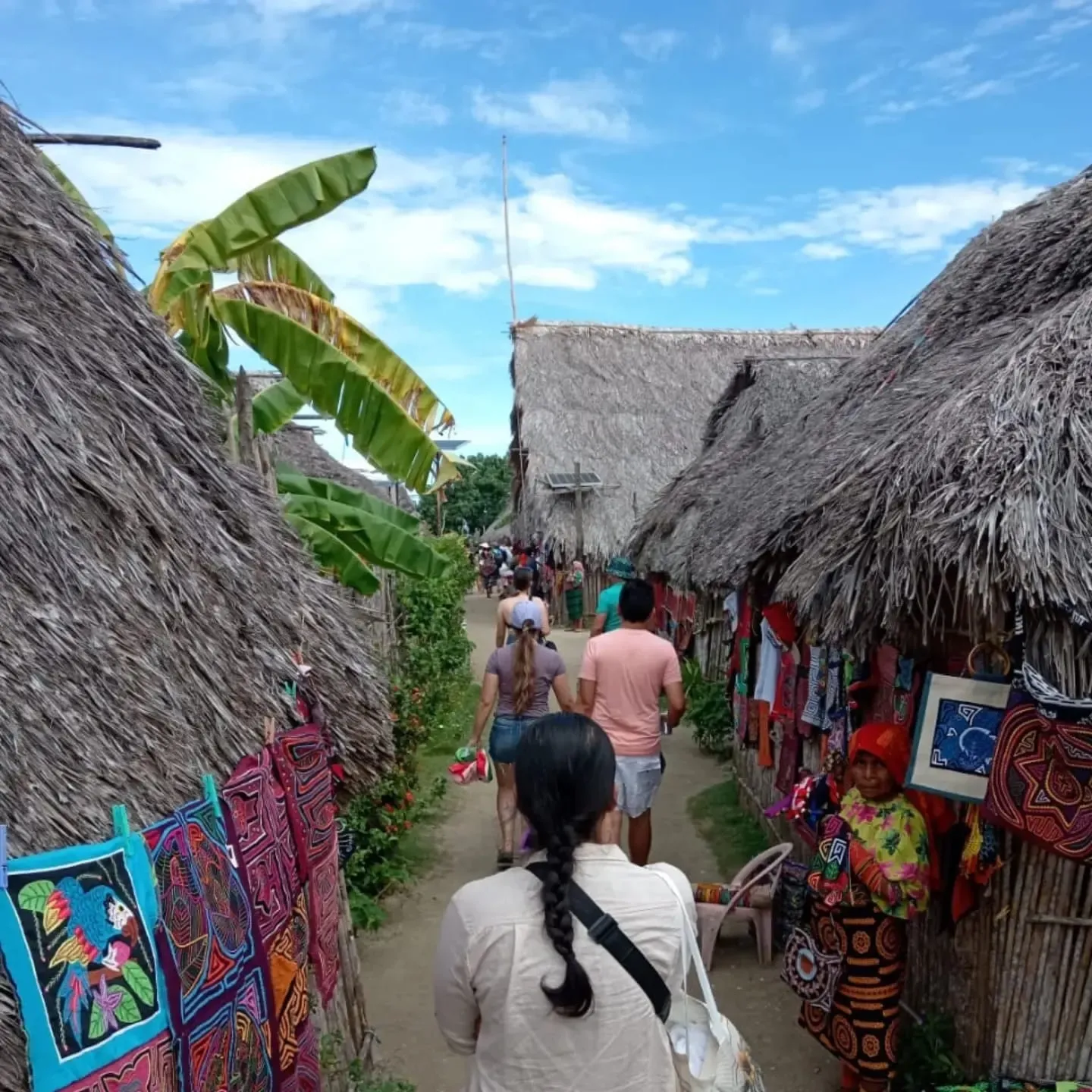 Visitantes caminan por una calle de tierra entre casas de techo de paja con molas colgadas, en una aldea de Guna Yala